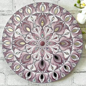 May include: A round mandala painted on a wooden surface with a purple, white, and silver color scheme. The mandala features intricate floral designs and geometric patterns.