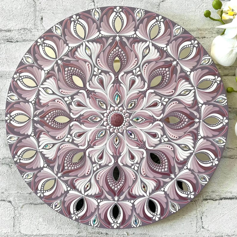 May include: A round mandala painted on a wooden surface with a purple, white, and silver color scheme. The mandala features intricate floral designs and geometric patterns.