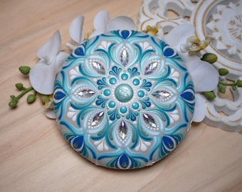 May include: A hand-painted mandala rock with a blue, white, and green design. The mandala features a central circle with a swirling pattern and is surrounded by intricate floral motifs.