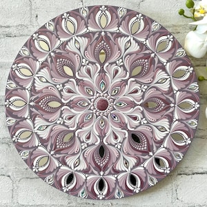 May include: A round mandala painted on a wooden surface with a purple and white design. The mandala features intricate details and a mirrored center.