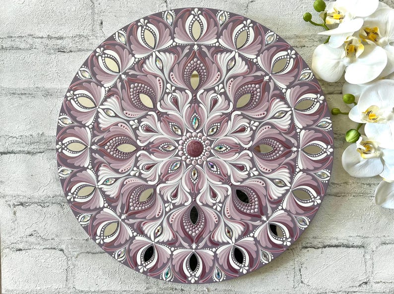 May include: A round mandala painted on a wooden surface with a purple and white design. The mandala features intricate details and a mirrored center.