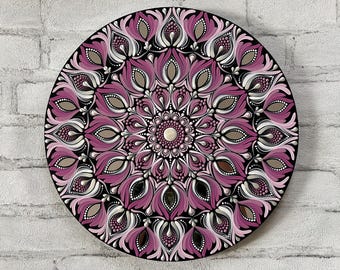 May include: A circular mandala painted on a wooden surface. The mandala features a repeating pattern of pink, white, and silver flowers and leaves on a black background.