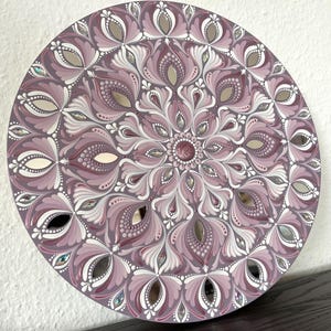 May include: A round mandala painted on a wooden board with a pink and white floral design. The mandala is decorated with small mirrored tiles.
