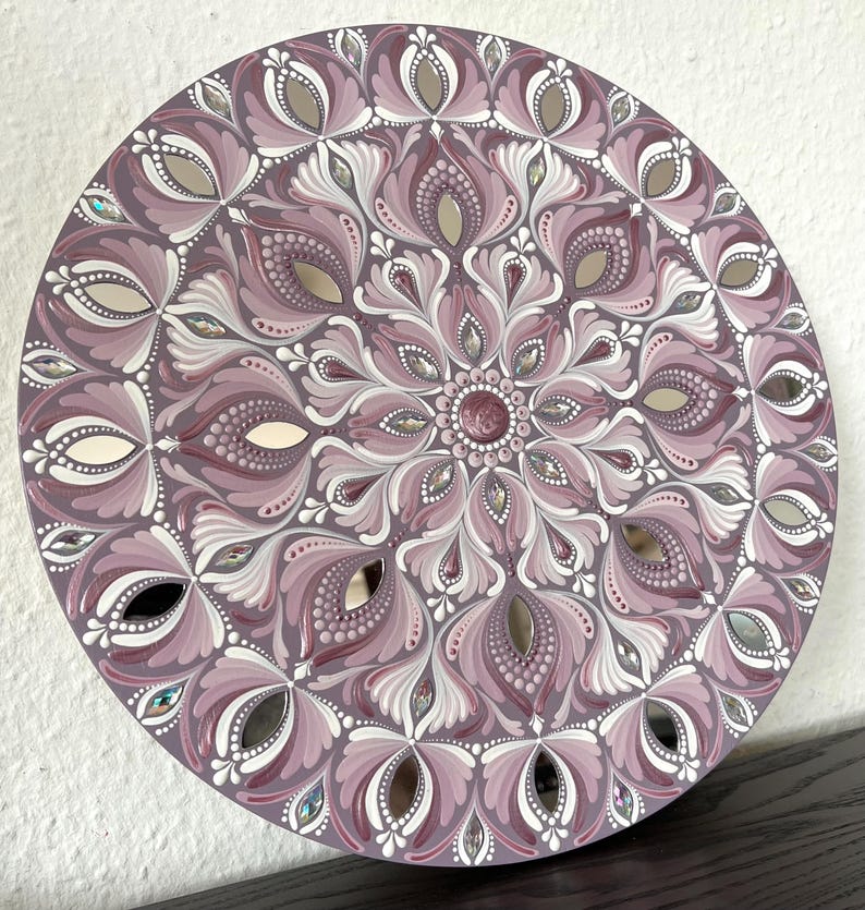 May include: A round mandala painted on a wooden board with a pink and white floral design. The mandala is decorated with small mirrored tiles.