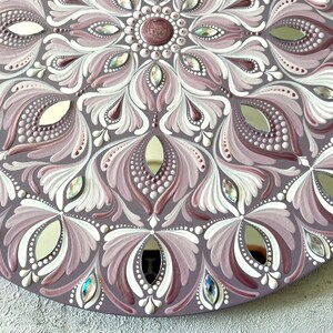 May include: A circular mandala painted on a wooden surface with a mirrored center. The mandala features a repeating pattern of white and pink floral designs with iridescent rhinestones.