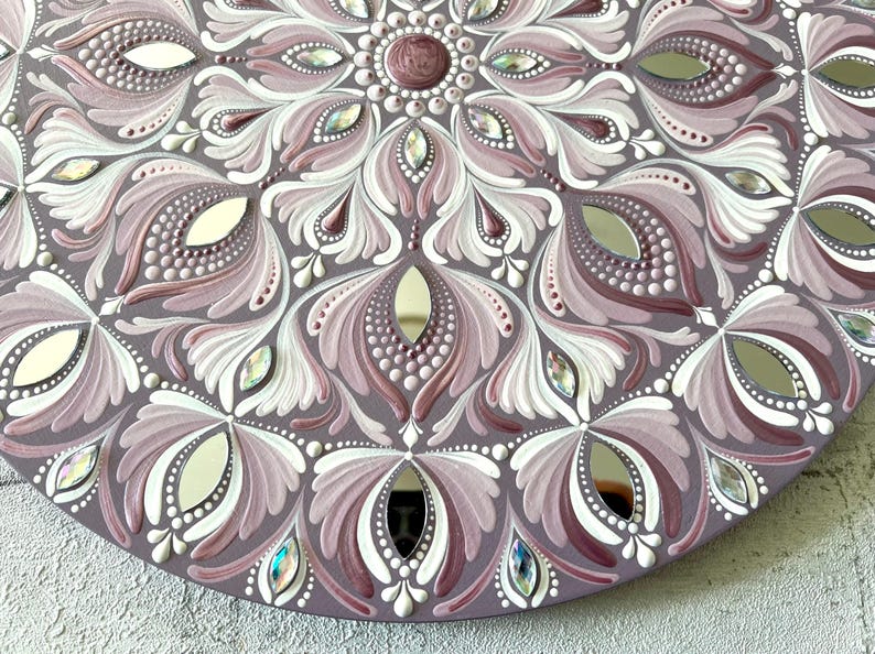 May include: A circular mandala painted on a wooden surface with a mirrored center. The mandala features a repeating pattern of white and pink floral designs with iridescent rhinestones.