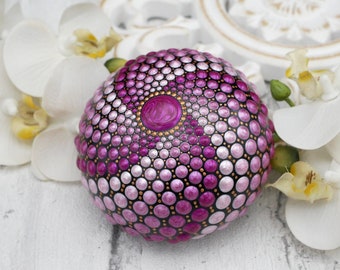 May include: A hand-painted rock with a mandala design in shades of pink, purple, and gold. The rock is surrounded by white orchids.