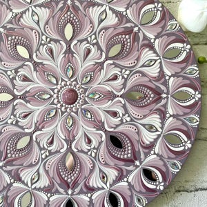 May include: A round mandala wall decor with a purple and white floral design. The mandala is painted with white and purple acrylic paint and decorated with rhinestones.