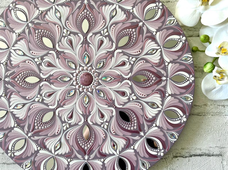May include: A round mandala wall decor with a purple and white floral design. The mandala is painted with white and purple acrylic paint and decorated with rhinestones.