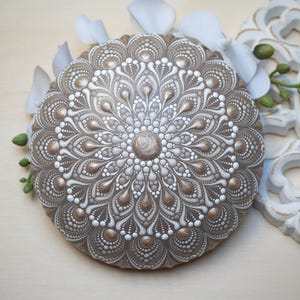 May include: A circular, hand-painted mandala stone in brown and white. The detailed design features a central point surrounded by patterns of dots and teardrop shapes. Decorative art.