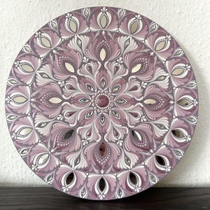 May include: A round mandala wall art piece with a pink and white design. The mandala is painted on a wooden surface and has a mirrored finish. The design features intricate details and a variety of shapes.