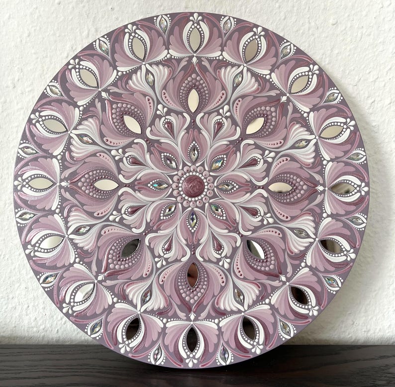 May include: A round mandala wall art piece with a pink and white design. The mandala is painted on a wooden surface and has a mirrored finish. The design features intricate details and a variety of shapes.