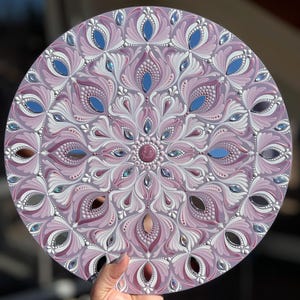 May include: A circular mandala painted on a wooden board with a pink, white, and silver color scheme. The mandala features a repeating pattern of floral designs and mirrored accents.