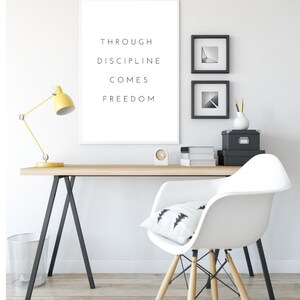 Pode incluir: Um escrit&oacute;rio em casa minimalista com uma mesa branca e uma cadeira branca. Uma impress&atilde;o emoldurada com o texto "Through discipline comes freedom" est&aacute; pendurada na parede. H&aacute; duas fotos emolduradas em preto e branco na parede acima da mesa.