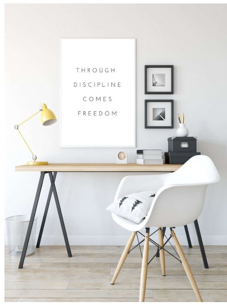 Pode incluir: Um escrit&oacute;rio em casa minimalista com uma mesa branca e uma cadeira branca. Uma impress&atilde;o emoldurada com o texto "Through discipline comes freedom" est&aacute; pendurada na parede. H&aacute; duas fotos emolduradas em preto e branco na parede acima da mesa.