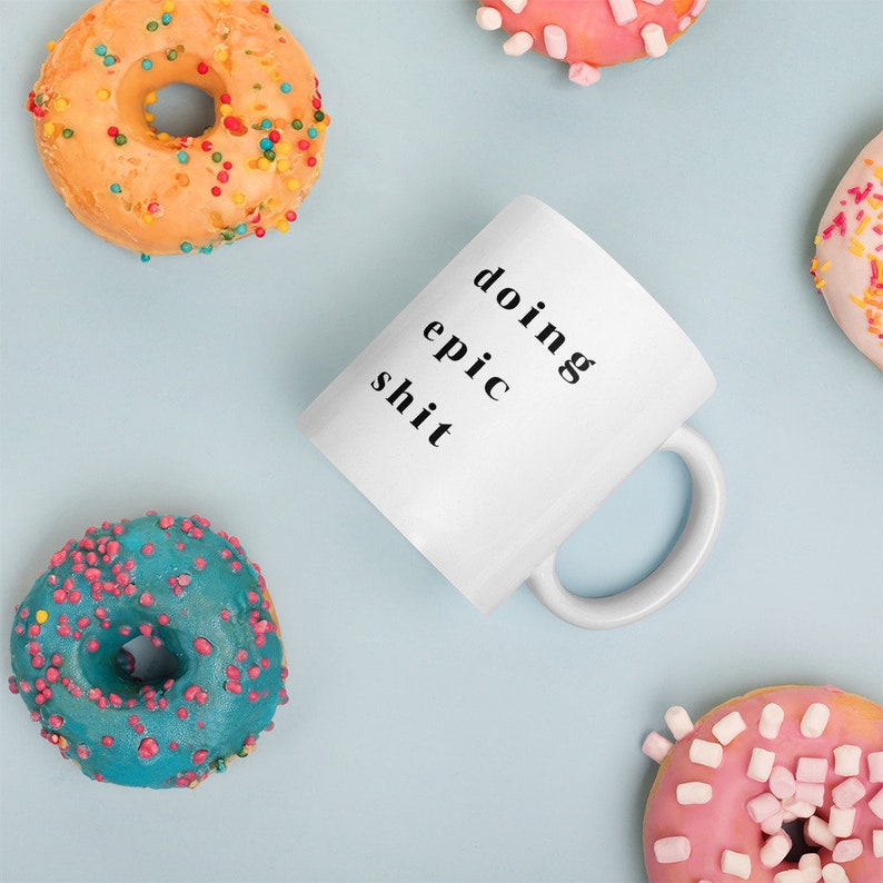 May include: White ceramic mug with black text that reads "doing epic shit". The mug is surrounded by colorful donuts with sprinkles.