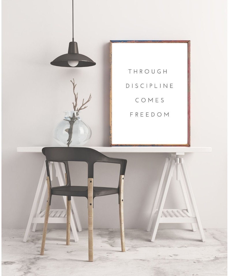 Pode incluir: Um p&ocirc;ster motivacional emoldurado em branco com o texto "Through discipline comes freedom" pendurado em uma parede branca acima de uma mesa branca com uma cadeira preta e cavaletes brancos.