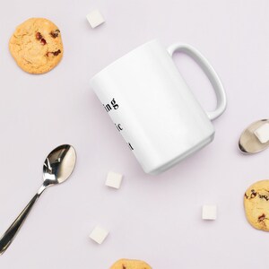 May include: White ceramic mug with black text that reads "Nothing is magic until you believe it". The mug is on a light purple surface with sugar cubes, cookies, and a silver spoon.