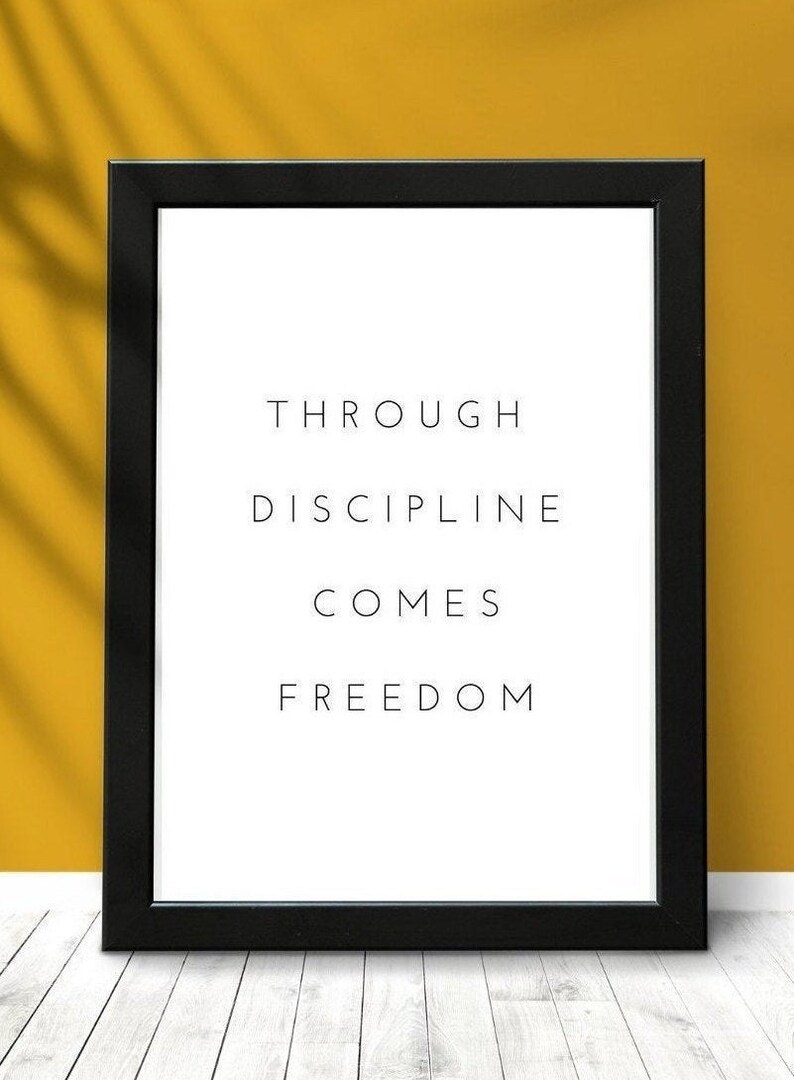 Pode incluir: Impress&atilde;o emoldurada em preto com o texto "Through discipline comes freedom" em fundo branco. A impress&atilde;o est&aacute; sobre uma superf&iacute;cie de madeira branca com uma parede amarela atr&aacute;s.