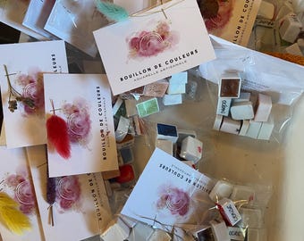 May include: Watercolor paint sets with small, square paint cakes in various colors. Each set includes a card with the text "BOUILLON DE COULEURS AQUARELLE ARTISANALE" and decorative elements. Some sets have dried flowers or feathers.