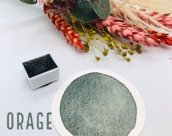 May include: A small, white pan with a black watercolor paint inside. The paint is a dark green color. The pan is sitting on a white surface. The word "ORAGE" is written in green letters below the pan.