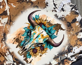 May include: A white long-sleeve shirt with a brown and black cow print pattern. The shirt features a graphic of a white bull skull with turquoise and yellow accents, surrounded by sunflowers.