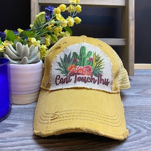 May include: A yellow distressed trucker hat with a patch featuring a cactus and the text "Can't Touch This".