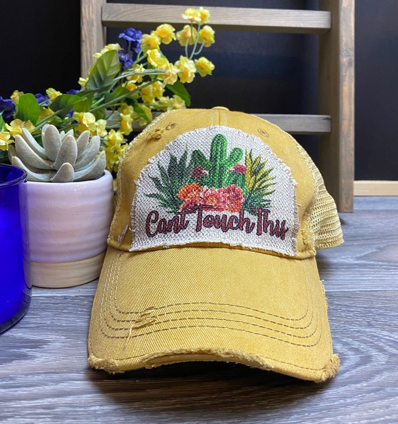 May include: A yellow distressed trucker hat with a patch featuring a cactus and the text "Can't Touch This".