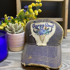 May include: A distressed denim baseball cap with a brown patch featuring a skull with floral accents and the text "Gypsy Soul".