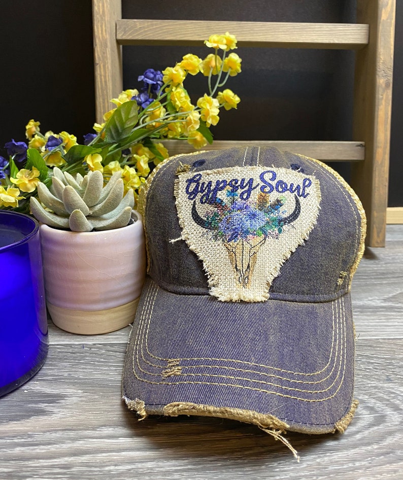 May include: A distressed denim baseball cap with a brown patch featuring a skull with floral accents and the text "Gypsy Soul".