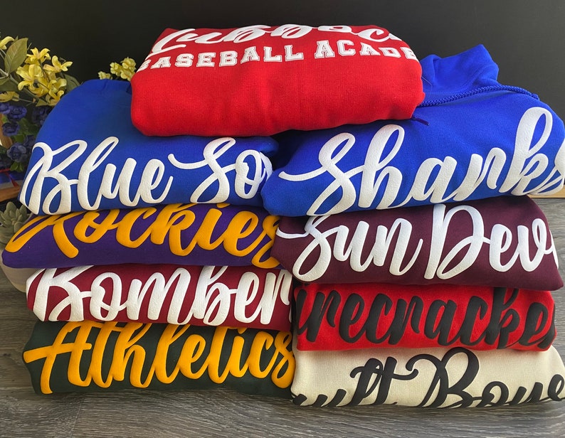 May include: A stack of ten hooded sweatshirts in various colors, each with a different team name printed in white or black cursive lettering. The names include "Blue Sox", "Shanks", "Rockies", "Sun Devils", "Bombers", "Firecrackers", "Athletics", and "Jet Boys".