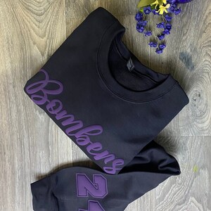 May include: Black sweatshirt with purple text that says "Bombers" and a purple number "24" on the sleeve.