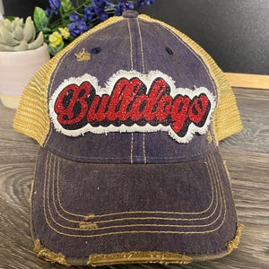 May include: A distressed navy blue baseball cap with a gold mesh back. The front features a white patch with frayed edges and the word Bulldogs in red glitter script with a black outline. The cap has visible stitching and worn fabric details.