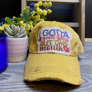 May include: A mustard yellow trucker hat with a distressed look. The hat has a patch on the front that says "I gotta good heart but this mouth" with a red lip print.