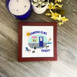 May include: A framed tile with a blue and white camper van illustration. The camper van has a paisley pattern and a blue door. The text "Camping is my therapy" is written in blue letters on the tile.