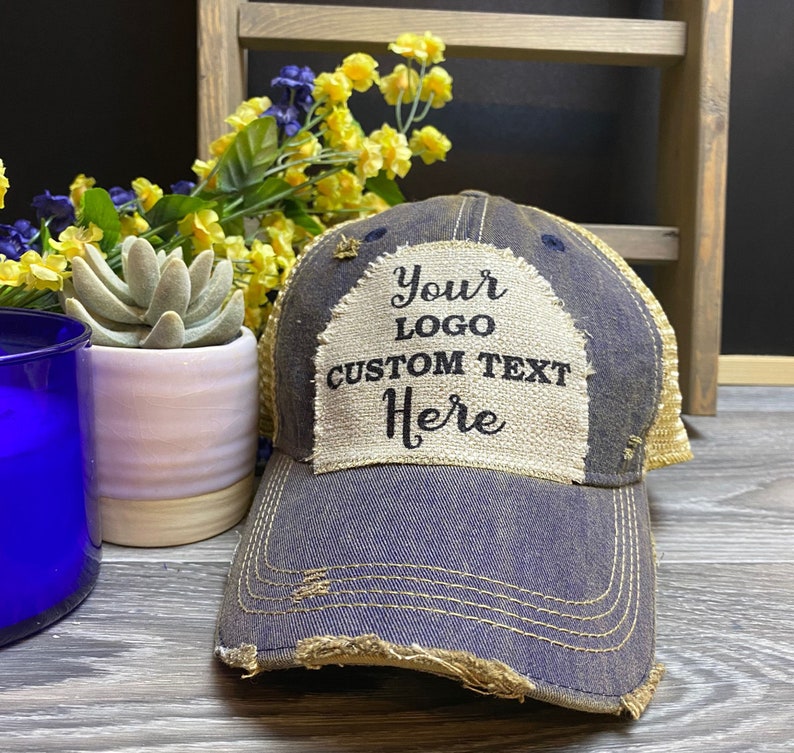 May include: A blue and beige distressed trucker hat with a beige patch that says "Your Logo Custom Text Here" in black text.