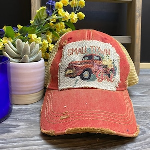 May include: Red and tan trucker hat with a distressed look. The hat has a patch on the front that says "Small Town Girl" and features a red pickup truck with flowers.