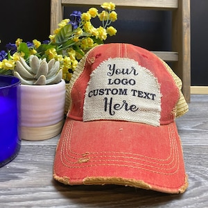 May include: Red and beige distressed trucker hat with a beige patch that says "Your Logo Custom Text Here" in black text.