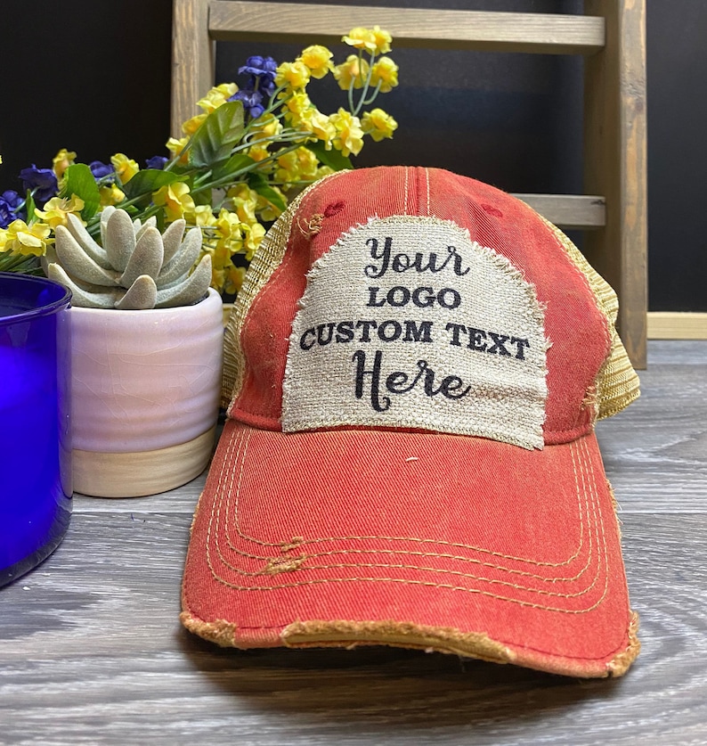 May include: Red and beige distressed trucker hat with a beige patch that says "Your Logo Custom Text Here" in black text.