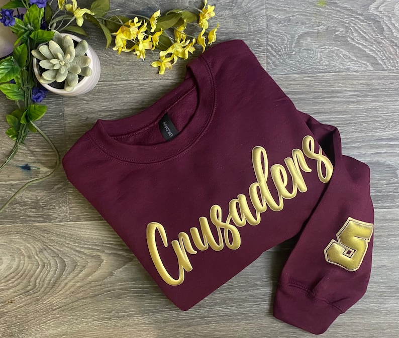 May include: Burgundy sweatshirt with gold lettering that says "Crusaders" and a gold number "5" on the sleeve.