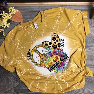 May include: Mustard yellow t-shirt with a tie-dye effect. The shirt features a peace sign, a leopard print heart, and a colorful sunflower design. Text includes "peace", "love", and "Hippie Soul".