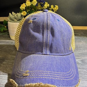 May include: A distressed purple baseball cap with a tan mesh back and brim. The cap features a curved brim, decorative stitching, and small star accents. The hat is designed with a worn, vintage aesthetic.