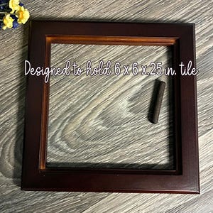 May include: A square, dark brown wooden frame designed to hold a 6 x 6 x 0.25 inch tile. The frame has a lighter brown inner border. The text "Designed to hold 6 x 6 x .25 in. tile" is written in cursive. A small, dark brown object is inside the frame.