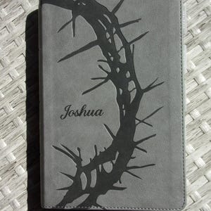 May include: Gray journal with a dark gray thorn design and the name "Joshua" in script. The journal has a ribbon bookmark and is on a woven surface.