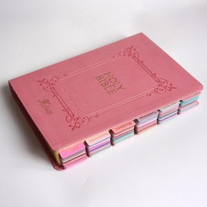 May include: A pink Holy Bible with gold lettering and decorative borders. The cover is embossed with the words "Holy Bible" and the name "Kira". The pages are separated by colorful tabs.