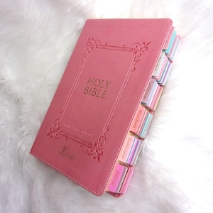 May include: A pink Holy Bible with gold lettering and decorative border. The book has labeled tabs in various pastel colors. The cover reads "Holy Bible" and "King James Version". The name "Kira" is also printed on the cover.