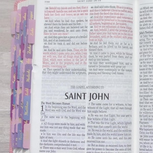 Pu&ograve; includere: Una Bibbia aperta che mostra la fine del Vangelo di Luca e l'inizio del Vangelo di San Giovanni. Le pagine presentano testo stampato in nero con passaggi evidenziati e una serie di linguette viola e rosa lungo il bordo sinistro.