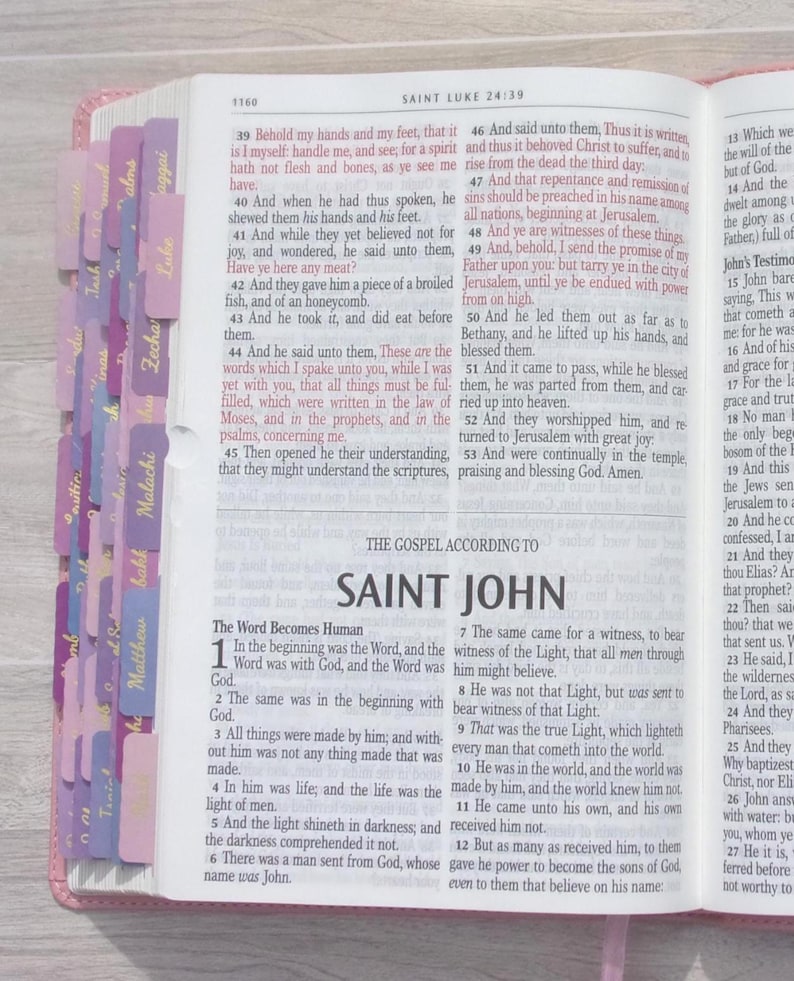 Pu&ograve; includere: Una Bibbia aperta che mostra la fine del Vangelo di Luca e l'inizio del Vangelo di San Giovanni. Le pagine presentano testo stampato in nero con passaggi evidenziati e una serie di linguette viola e rosa lungo il bordo sinistro.