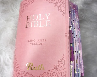 May include: A pink Holy Bible, King James Version, with gold lettering and decorative corner designs. The book is open, revealing color-coded page tabs. The word "Ruth" is printed in gold on the cover.