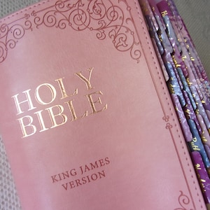 Pu&ograve; includere: Una Bibbia color rosa polvere, versione King James, con scritte in lamina d'oro sulla copertina. La copertina presenta un bordo decorativo a volute in rilievo. Il lato del libro include diverse linguette indice con motivi viola e lavanda.